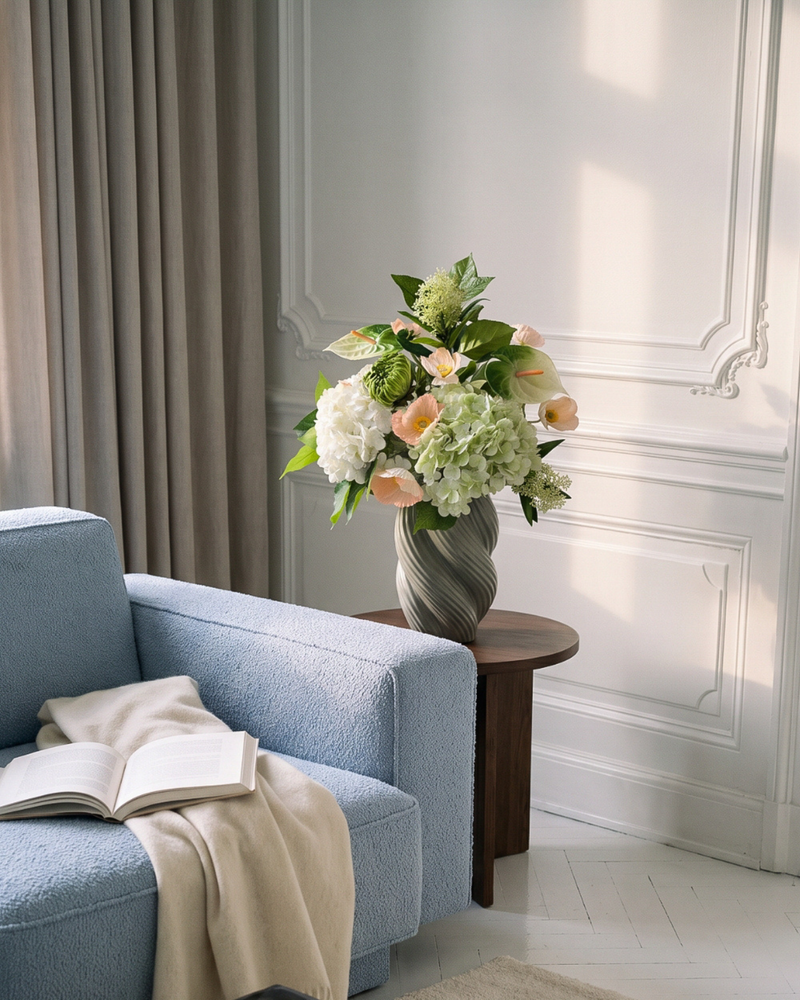 Lyserød, hvid og grøn kunstig buket med hortensia, anthurium og valmuer i en snoet keramikvase, placeret på et rundt mørkt træsidebord i en klassisk københavnerlejlighed med hvide vægpaneler og stuk. Varmt eftermiddagslys falder skråt ind over blomsterne. En lyseblå bouclé-sofa med en opslået bog og et cremefarvet plaid ses i forgrunden.
