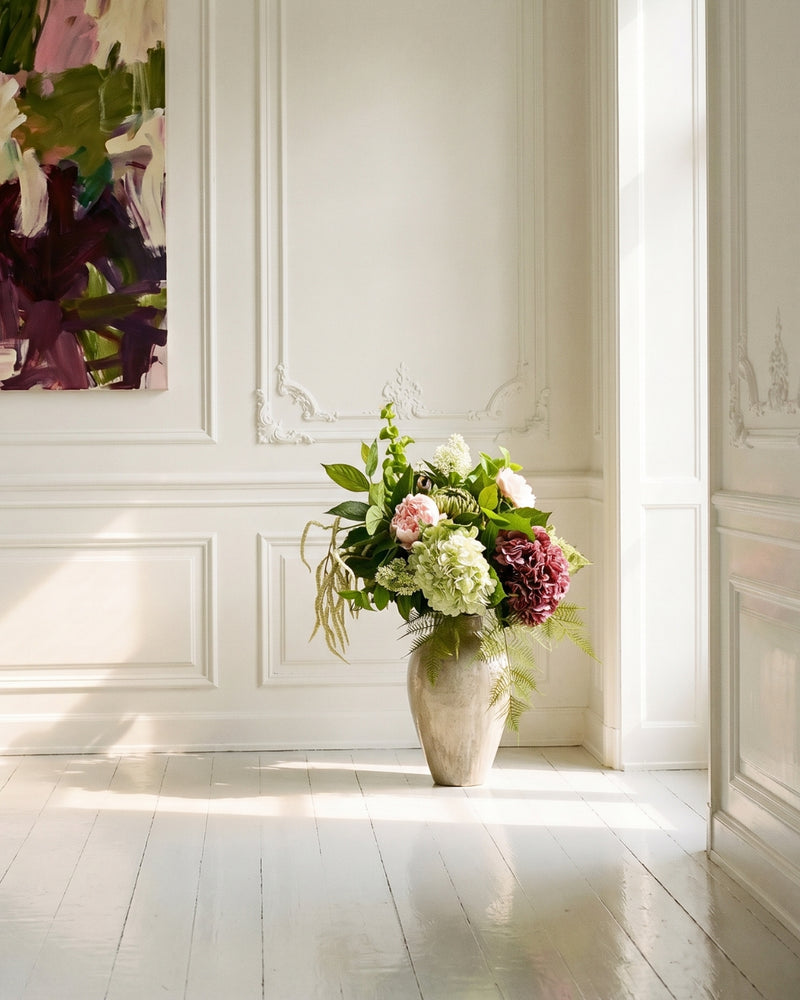 Kunstig Bloomgaard-buket med lyserøde pæoner, grøn hortensia, mørk dahlia, bregner og amaranthus i en stenvase med struktur, placeret på et hvidmalet trægulv i en solbeskinnet københavnerlejlighed med stukvægge og et abstrakt maleri i grønne og bordeaux toner