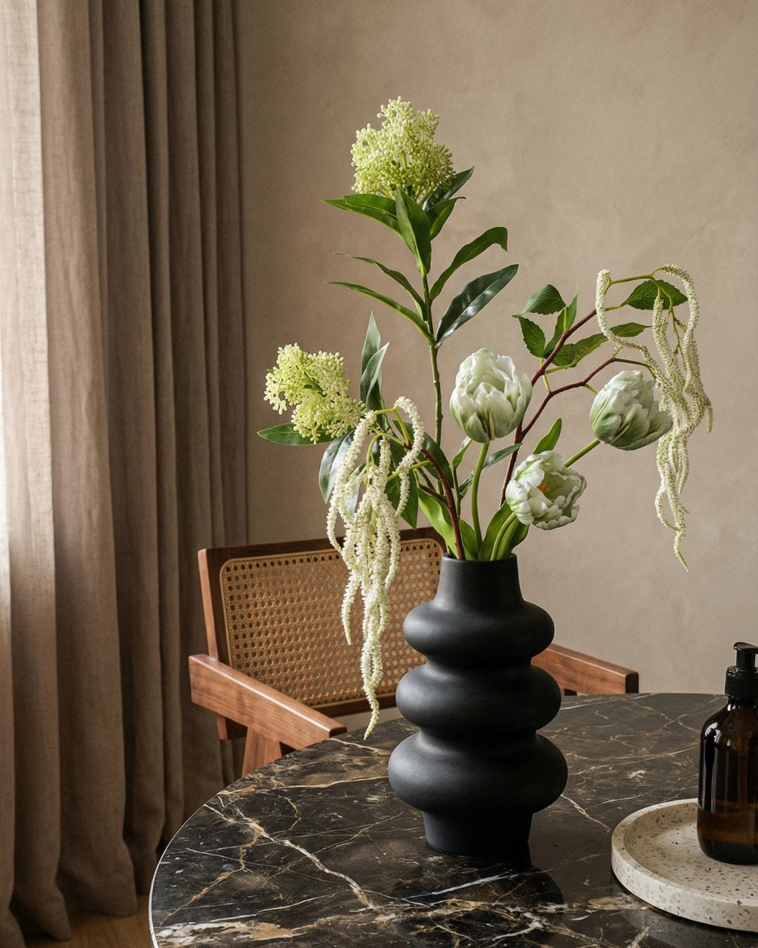 Grøn og hvid blomsterbuket i en sort vase placeret på et mørkt, rundt marmorbord i hjørnet af et rum