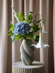 En elegant og luftig buket, hvor den blå hortensia danner et blødt centrum omgivet af friske grønne blade og fine hvide stramonium og rævehale. Sammensætningen balancerer det frodige og det lette på smukkeste vis og skaber et harmonisk og naturligt udtryk. Perfekt som en dekorativ detalje, der bringer friskhed og ro til både hjemmet og kontoret.