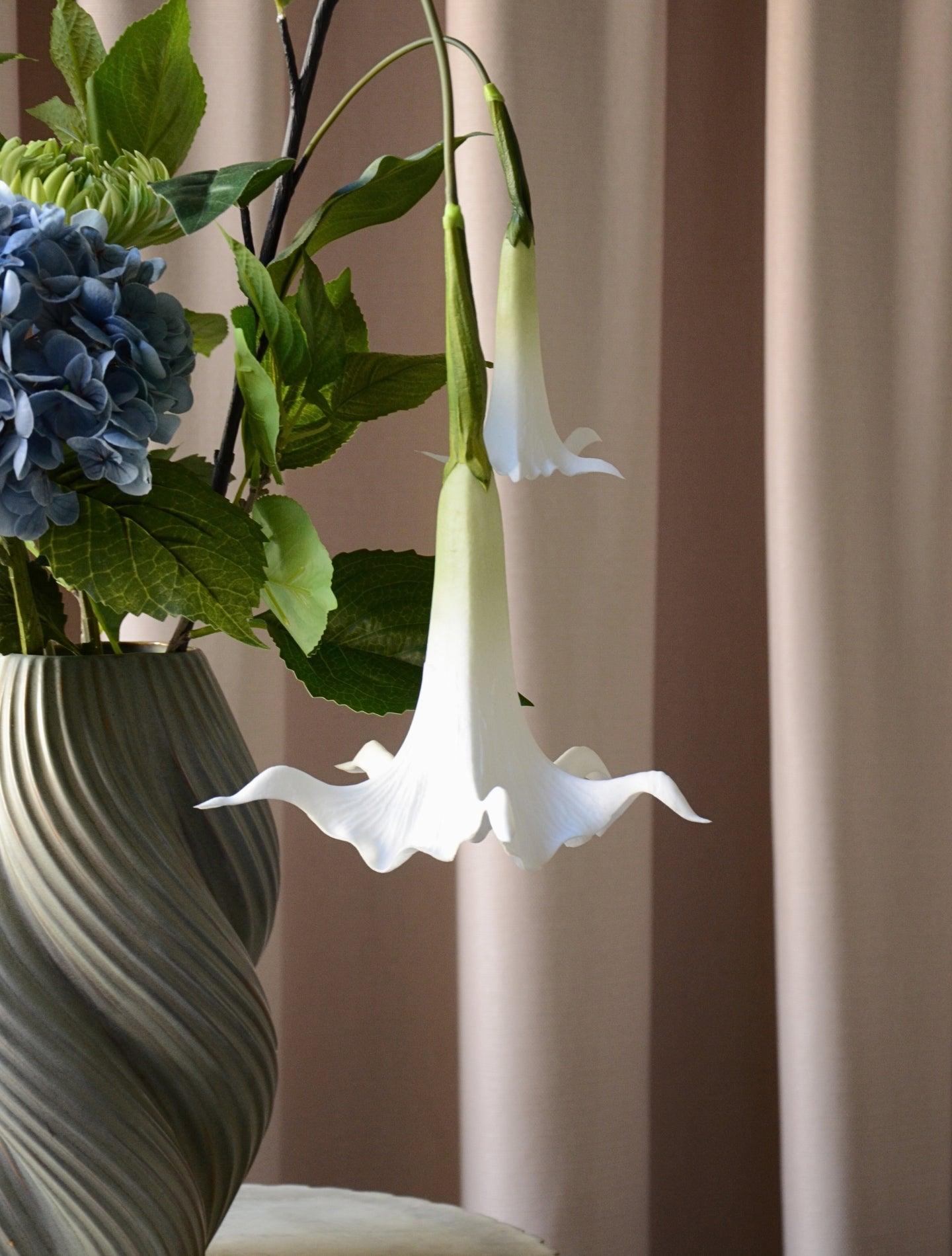 En elegant og luftig buket, hvor den blå hortensia danner et blødt centrum omgivet af friske grønne blade og fine hvide stramonium og rævehale. Sammensætningen balancerer det frodige og det lette på smukkeste vis og skaber et harmonisk og naturligt udtryk. Perfekt som en dekorativ detalje, der bringer friskhed og ro til både hjemmet og kontoret.