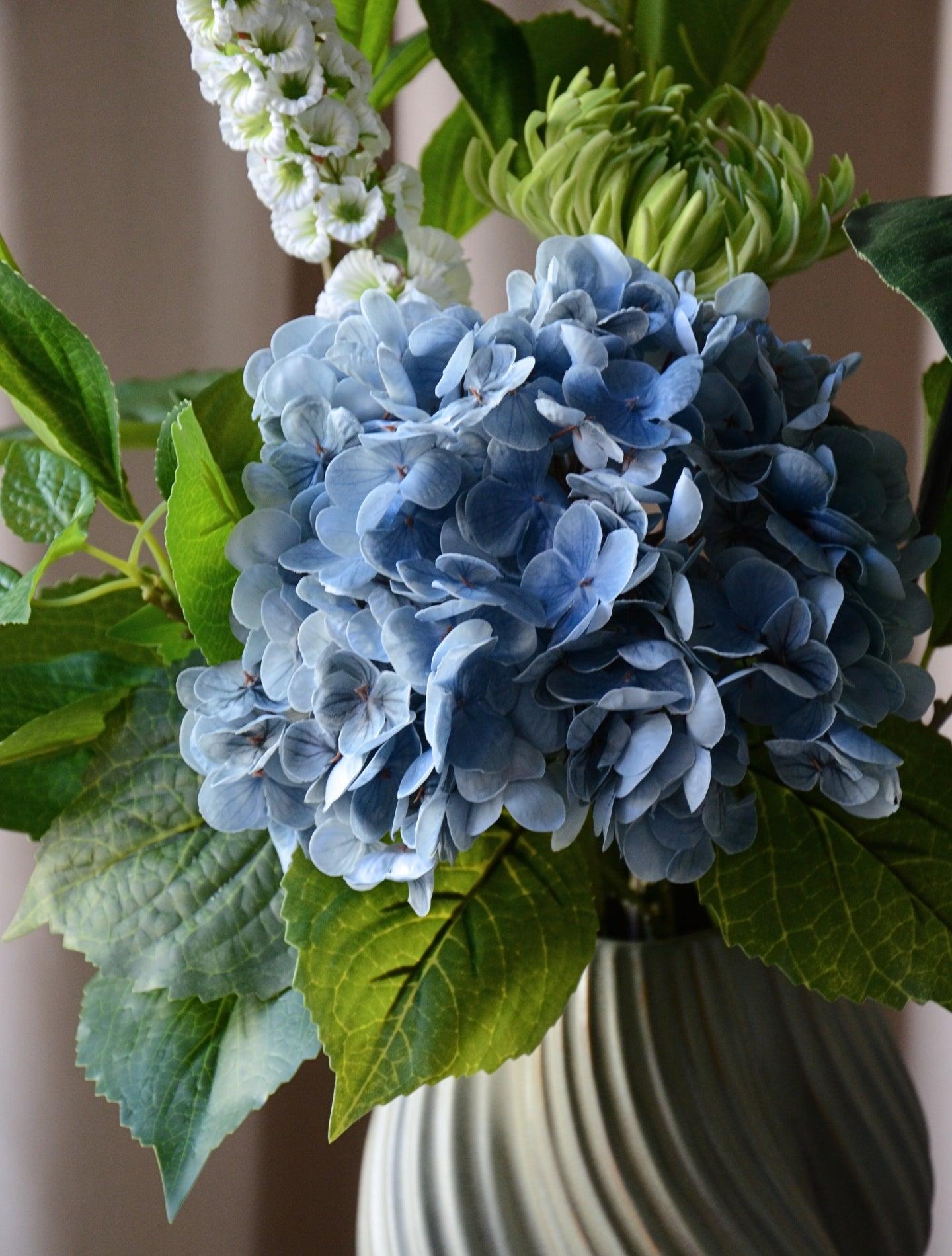 En elegant og luftig buket, hvor den blå hortensia danner et blødt centrum omgivet af friske grønne blade og fine hvide stramonium og rævehale. Sammensætningen balancerer det frodige og det lette på smukkeste vis og skaber et harmonisk og naturligt udtryk. Perfekt som en dekorativ detalje, der bringer friskhed og ro til både hjemmet og kontoret.