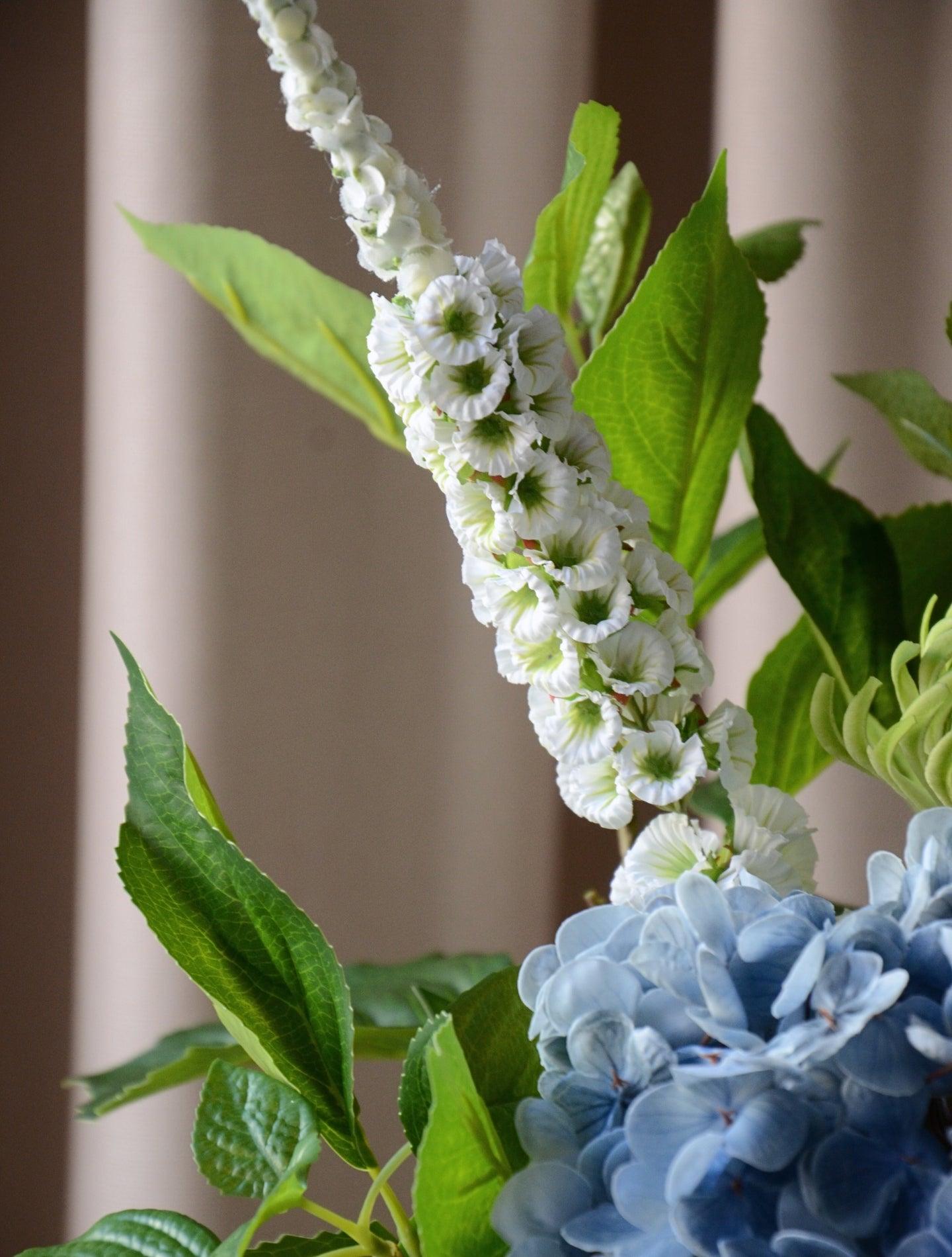 En elegant og luftig buket, hvor den blå hortensia danner et blødt centrum omgivet af friske grønne blade og fine hvide stramonium og rævehale. Sammensætningen balancerer det frodige og det lette på smukkeste vis og skaber et harmonisk og naturligt udtryk. Perfekt som en dekorativ detalje, der bringer friskhed og ro til både hjemmet og kontoret.