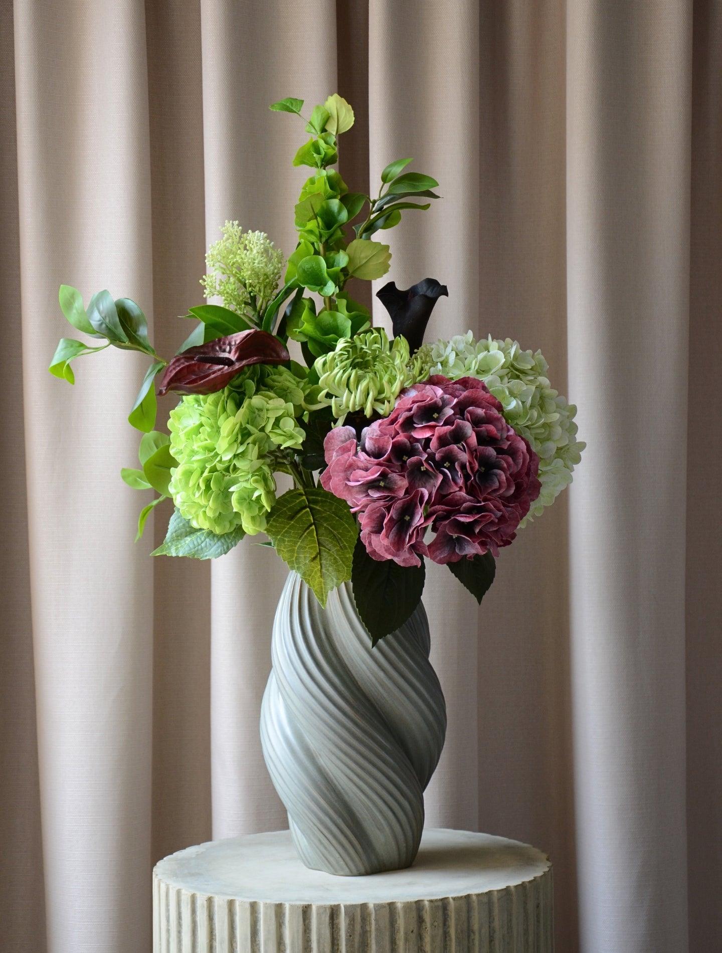 En elegant evighedsbuket, hvor mørk bordeaux møder friske limegrønne og støvede grønne nuancer i en smuk sammensætning. De fyldige hortensiahoveder, den karakterfulde calla lilje og anthurium og de lette grønne detaljer skaber et balanceret udtryk med både kant og finesse. En buket, der emmer af moderne luksus – ideel som dekorativ centerpiece i et stilfuldt hjem.