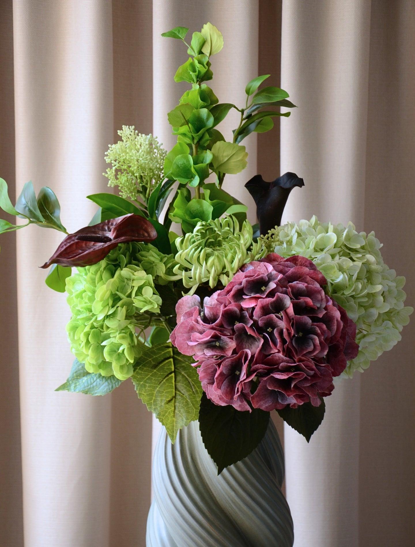 En elegant evighedsbuket, hvor mørk bordeaux møder friske limegrønne og støvede grønne nuancer i en smuk sammensætning. De fyldige hortensiahoveder, den karakterfulde calla lilje og anthurium og de lette grønne detaljer skaber et balanceret udtryk med både kant og finesse. En buket, der emmer af moderne luksus – ideel som dekorativ centerpiece i et stilfuldt hjem.