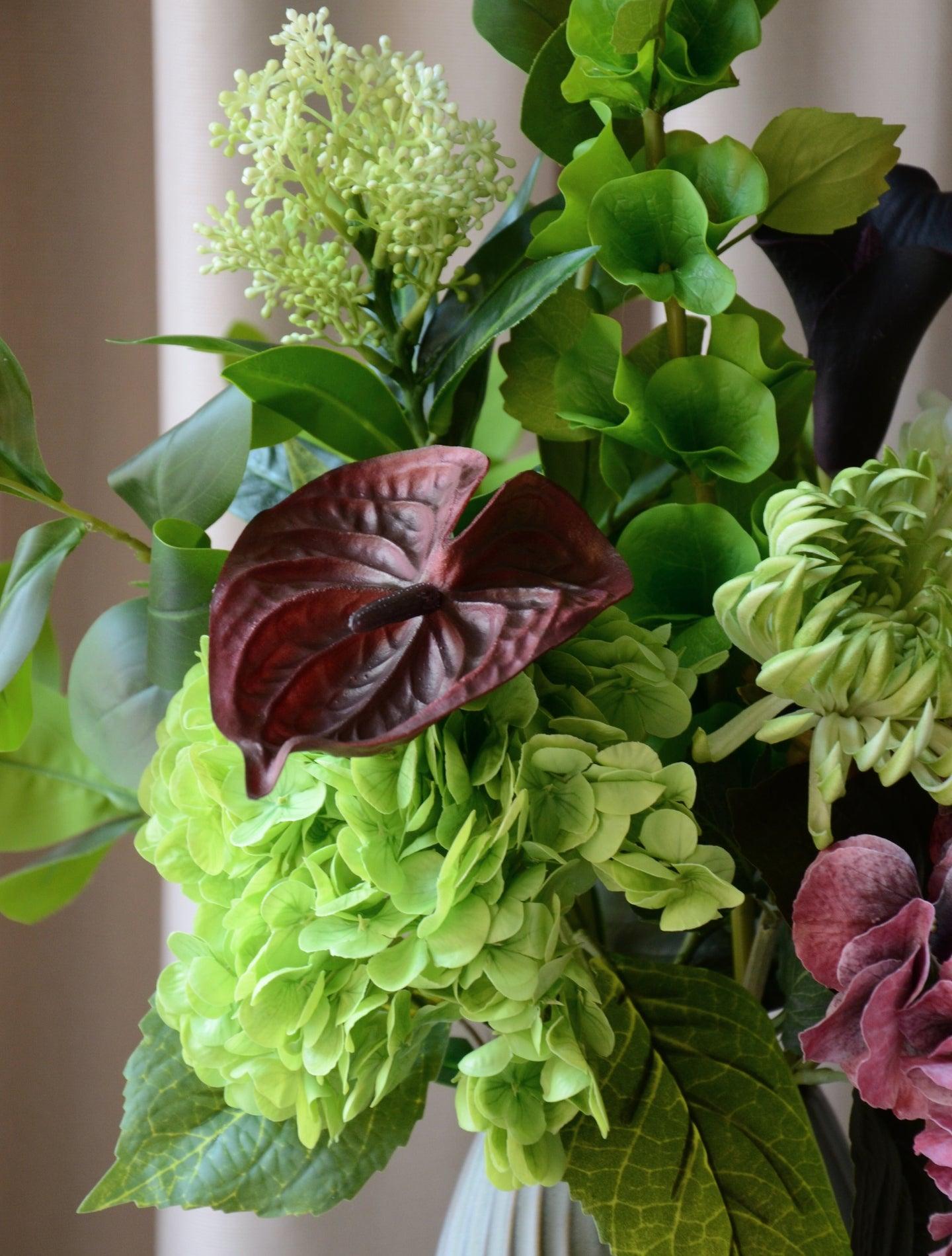 En elegant evighedsbuket, hvor mørk bordeaux møder friske limegrønne og støvede grønne nuancer i en smuk sammensætning. De fyldige hortensiahoveder, den karakterfulde calla lilje og anthurium og de lette grønne detaljer skaber et balanceret udtryk med både kant og finesse. En buket, der emmer af moderne luksus – ideel som dekorativ centerpiece i et stilfuldt hjem.
