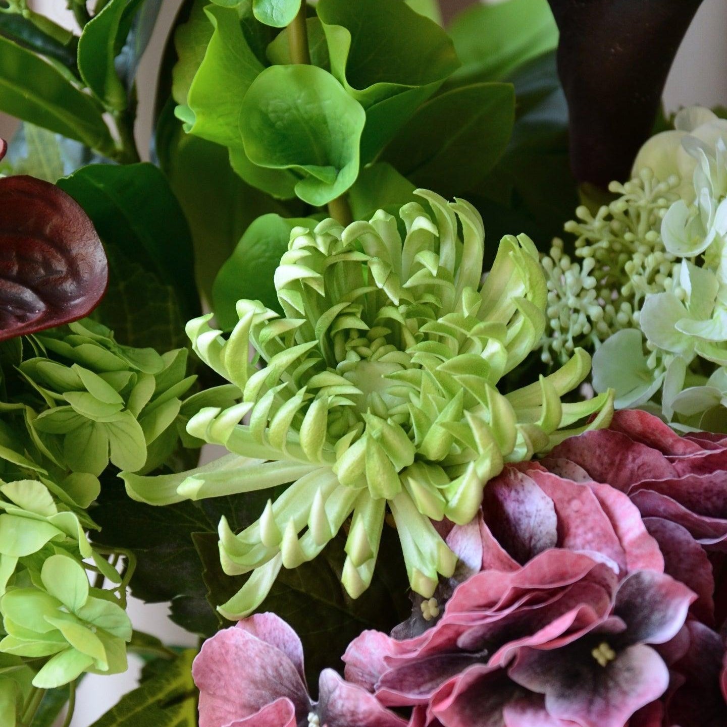 En elegant evighedsbuket, hvor mørk bordeaux møder friske limegrønne og støvede grønne nuancer i en smuk sammensætning. De fyldige hortensiahoveder, den karakterfulde calla lilje og anthurium og de lette grønne detaljer skaber et balanceret udtryk med både kant og finesse. En buket, der emmer af moderne luksus – ideel som dekorativ centerpiece i et stilfuldt hjem.