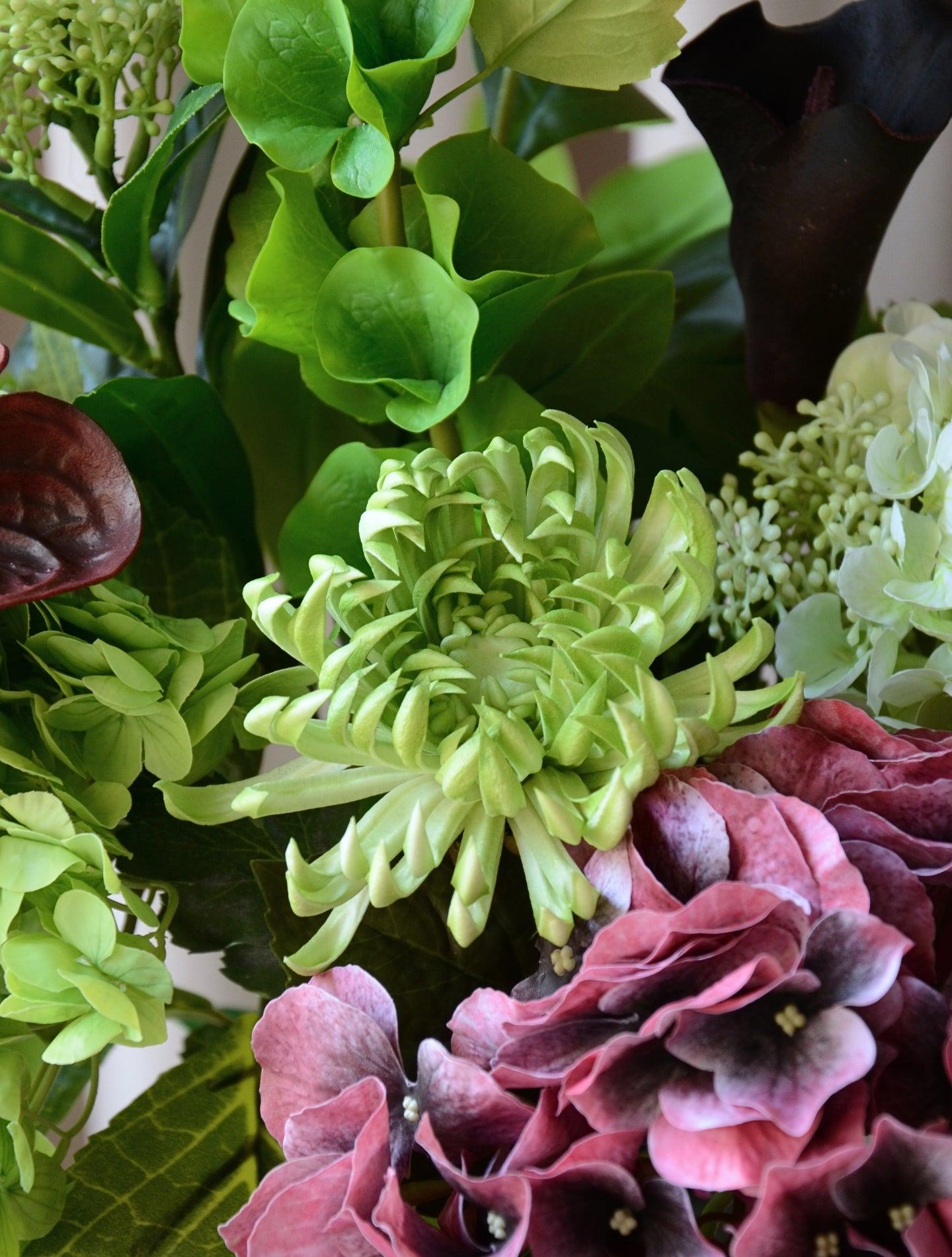 En elegant evighedsbuket, hvor mørk bordeaux møder friske limegrønne og støvede grønne nuancer i en smuk sammensætning. De fyldige hortensiahoveder, den karakterfulde calla lilje og anthurium og de lette grønne detaljer skaber et balanceret udtryk med både kant og finesse. En buket, der emmer af moderne luksus – ideel som dekorativ centerpiece i et stilfuldt hjem.