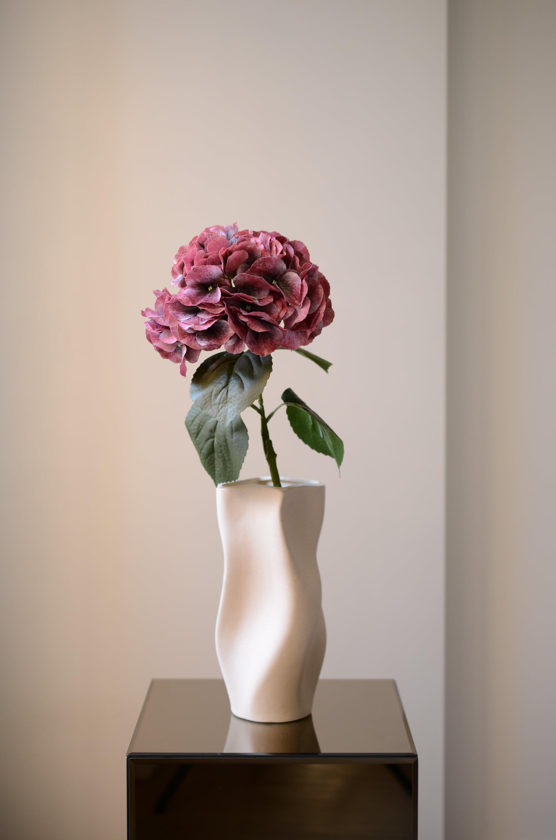 Denne kunstige hortensia i smukke, mørkrøde nuancer udstråler varme og eksklusivitet. Det fyldige blomsterhoved har en rig farvedybde med subtile nuancer, der skaber et levende og naturtro udtryk. De mørkegrønne blade langs stilken tilfører kontrast og detaljer til blomstens realistiske design. Perfekt som et elegant statement i en vase eller som en del af en evighedsbuket, der bevarer sin skønhed år efter år.