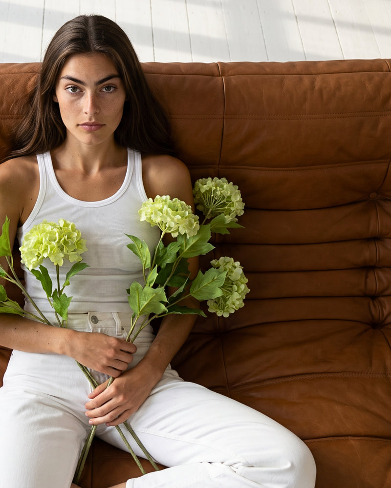 Kvinde i hvid tanktop og hvide jeans sidder på en cognacfarvet Togo-sofa med en håndfuld kunstige grønne snebolle-stilke