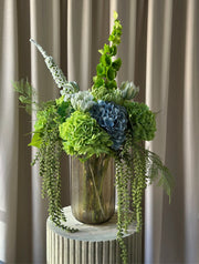 En stor, imponerende buket, hvor grønne nuancer smelter sammen med kølige blå toner i en elegant og luksuriøs komposition. Buketten emmer af eksklusivitet, ro og styrke – ideel som centerpiece eller som en markant, betagende dekoration i hjemmet eller på kontoret.

Buketten er bygget op af store, silkebløde hortensiahoveder i grøn og blå, omgivet af elegante tulipaner, chrysanthemum og karakterfulde foxtail og bells of ireland, der tilfører buketten en skulpturel lethed, mens de luftige bregner og hængende 