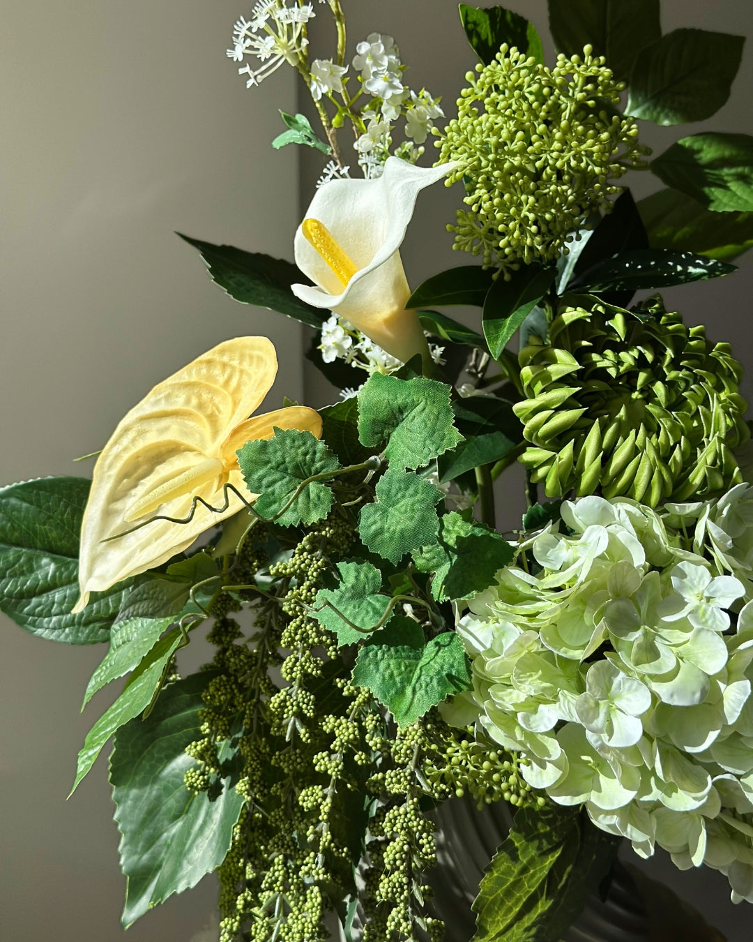 Green Loggia artificial bouquet with a light green hydrangea, a green cactus dahlia, a white calla lily and butter yellow anthurium in a grey-green twisted vase, with long green amaranthus trailing down onto a fluted stone pedestal