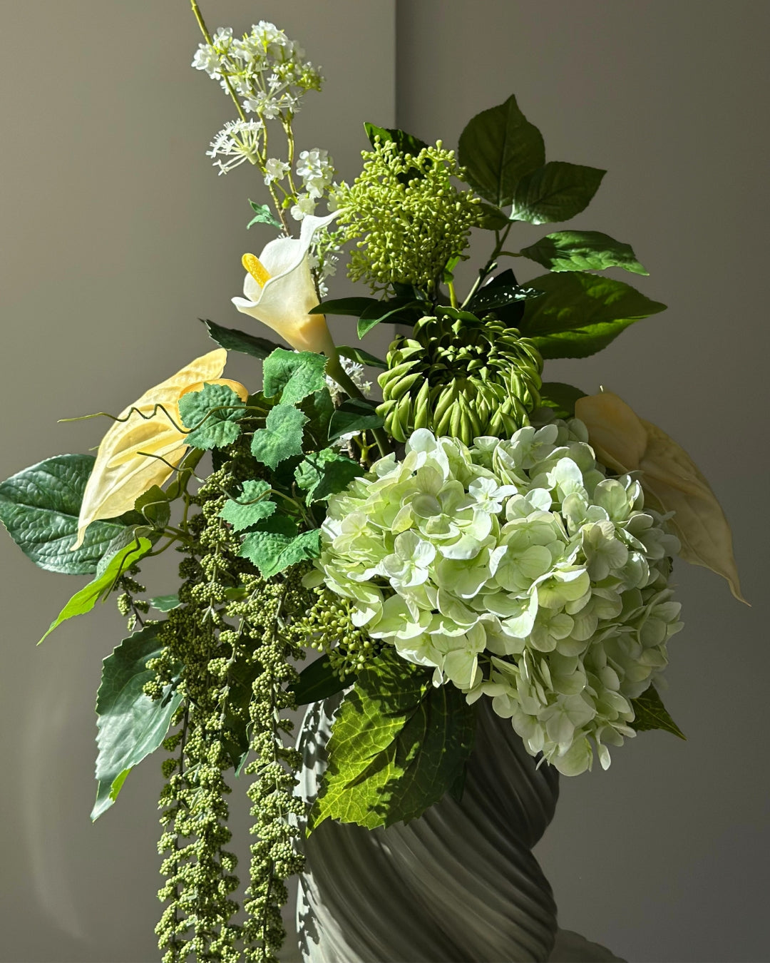Green Loggia artificial bouquet with a light green hydrangea, a green cactus dahlia, a white calla lily and butter yellow anthurium in a grey-green twisted vase, with long green amaranthus trailing down onto a fluted stone pedestal