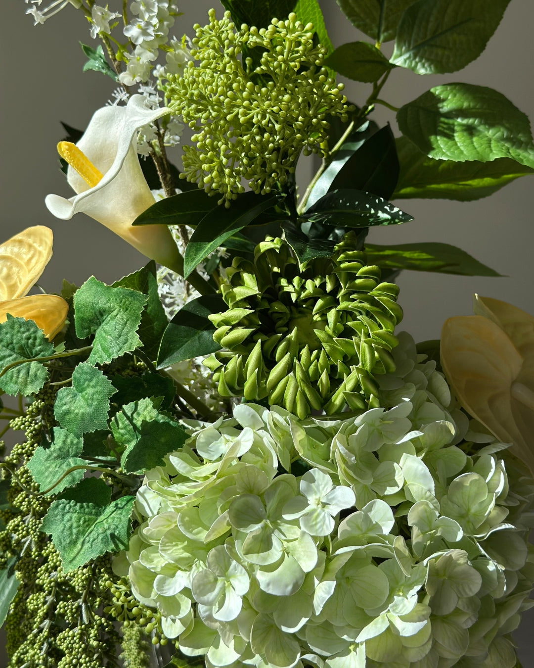Green Loggia artificial bouquet with a light green hydrangea, a green cactus dahlia, a white calla lily and butter yellow anthurium in a grey-green twisted vase, with long green amaranthus trailing down onto a fluted stone pedestal