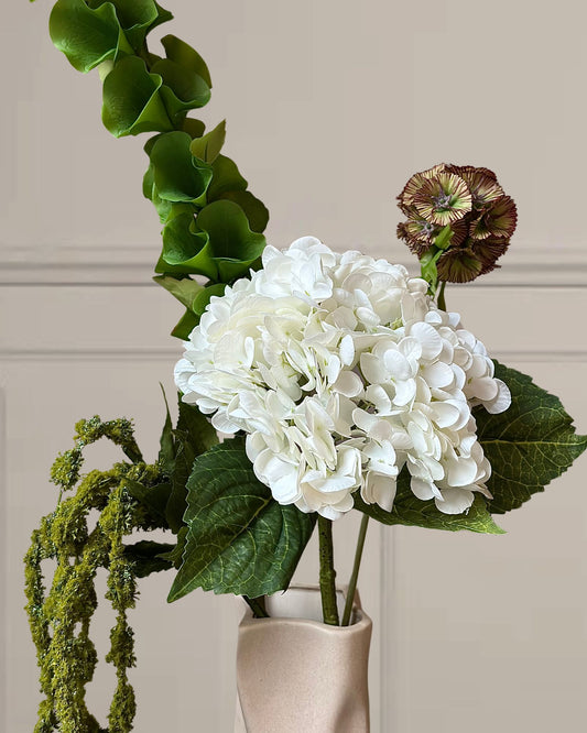 Denne smukke og enkle kunstige buket kombinerer tidløs elegance med naturlige farvetoner. En fyldig hvid hortensia danner bukettens står i kontrast til den karakteristiske grønne Bells of Ireland, Rævehalen og den smukke brune Skabiose.

Farvepaletten i hvid, grøn og brun skaber et roligt og afbalanceret udtryk, der passer perfekt til enhver indretning – fra det minimalistiske til det rustikke.