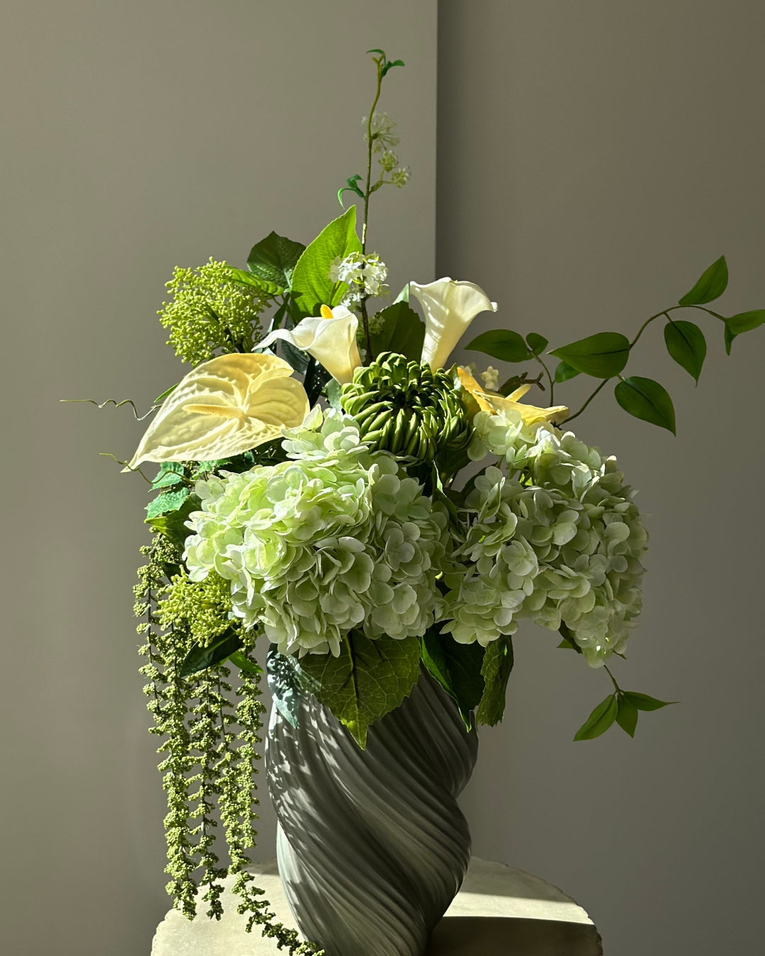 Green Atrium kunstig buket med grønne hortensiaer, hvide calla, grøn krysantemum og hængende amaranthus i en grågrøn snoet vase på en kannelleret stenpiedestal