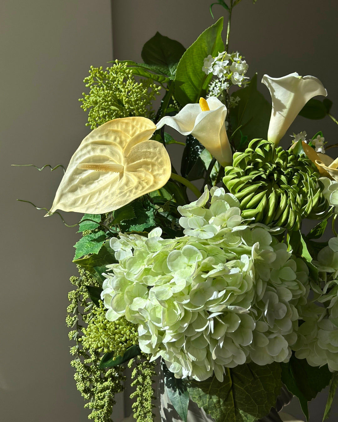Green Atrium kunstig buket med grønne hortensiaer, hvide calla, grøn krysantemum og hængende amaranthus i en grågrøn snoet vase på en kannelleret stenpiedestal