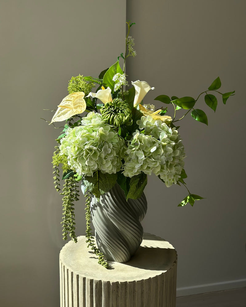 Green Atrium kunstig buket med grønne hortensiaer, hvide calla, grøn krysantemum og hængende amaranthus i en grågrøn snoet vase på en kannelleret stenpiedestal