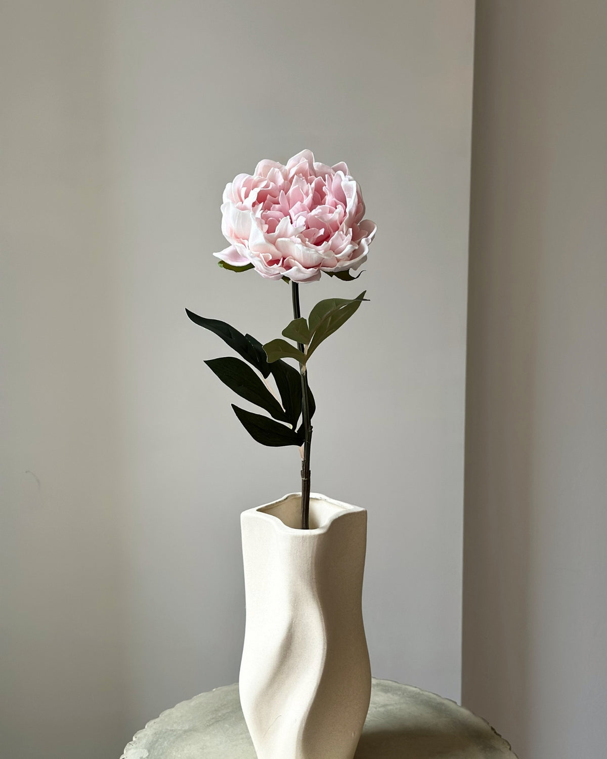 Stor, kunstig lyseroed pæon blomst med mørkegrønne blade på stilken. Den er placeret i en sandfarvet vase