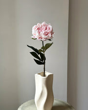 Stor, kunstig lyseroed pæon blomst med mørkegrønne blade på stilken. Den er placeret i en sandfarvet vase