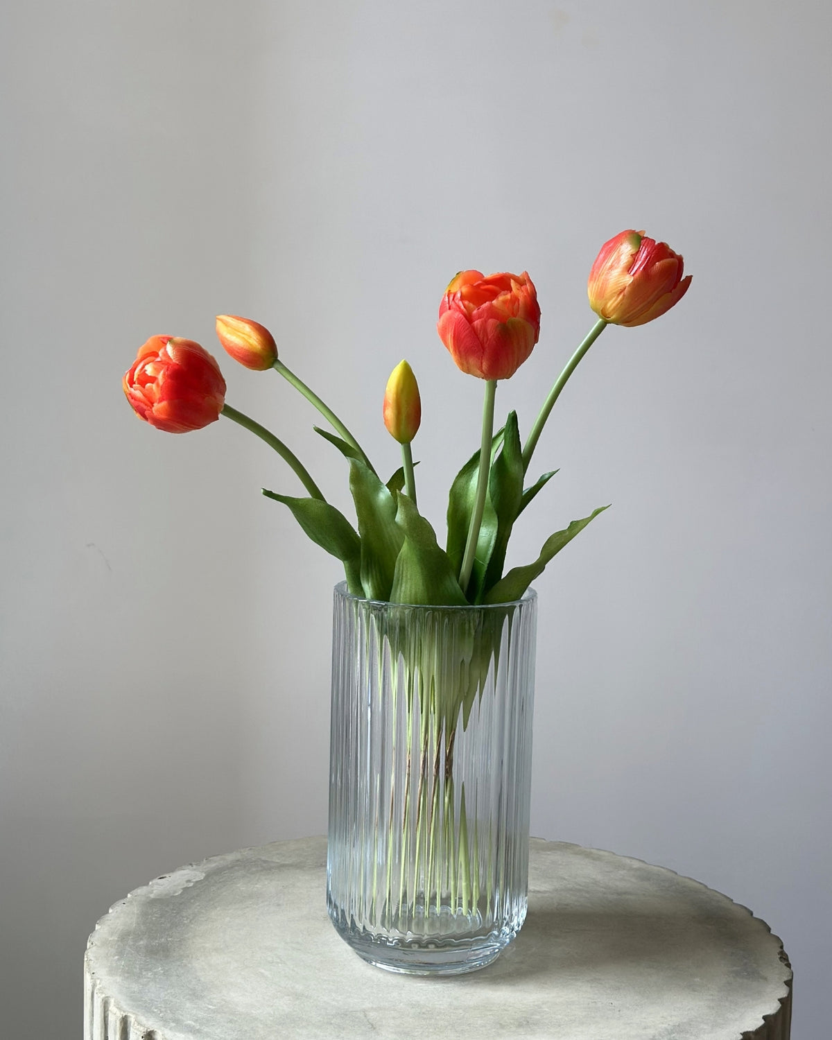 5 orange tulipaner i en glasvase på en cementpiedestal