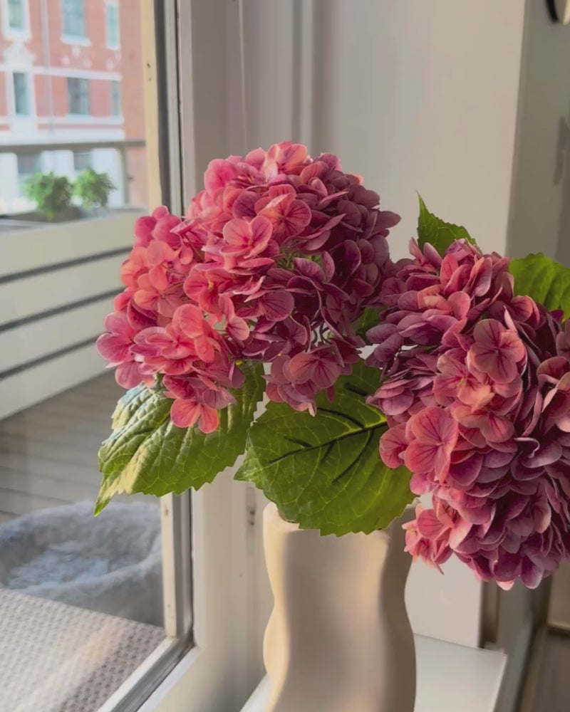 Den kunstige pink hortensia er en iøjnefaldende blomst, der tilføjer et romantisk og levende element til enhver buket eller dekoration. Med sine fyldige blomsterhoveder i smukke, varme nuancer skaber den en farverig og indbydende atmosfære. De detaljerede blomster og de grønne blade giver et naturligt og realistisk udtryk, mens den langvarige kvalitet gør den til en vedligeholdelsesfri løsning.

Stilken måler 49 cm.