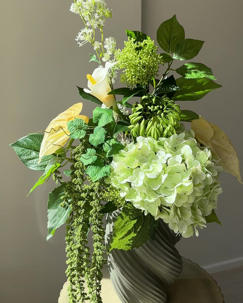 Green Loggia artificial bouquet with a light green hydrangea, a green cactus dahlia, a white calla lily and butter yellow anthurium in a grey-green twisted vase, with long green amaranthus trailing down onto a fluted stone pedestal