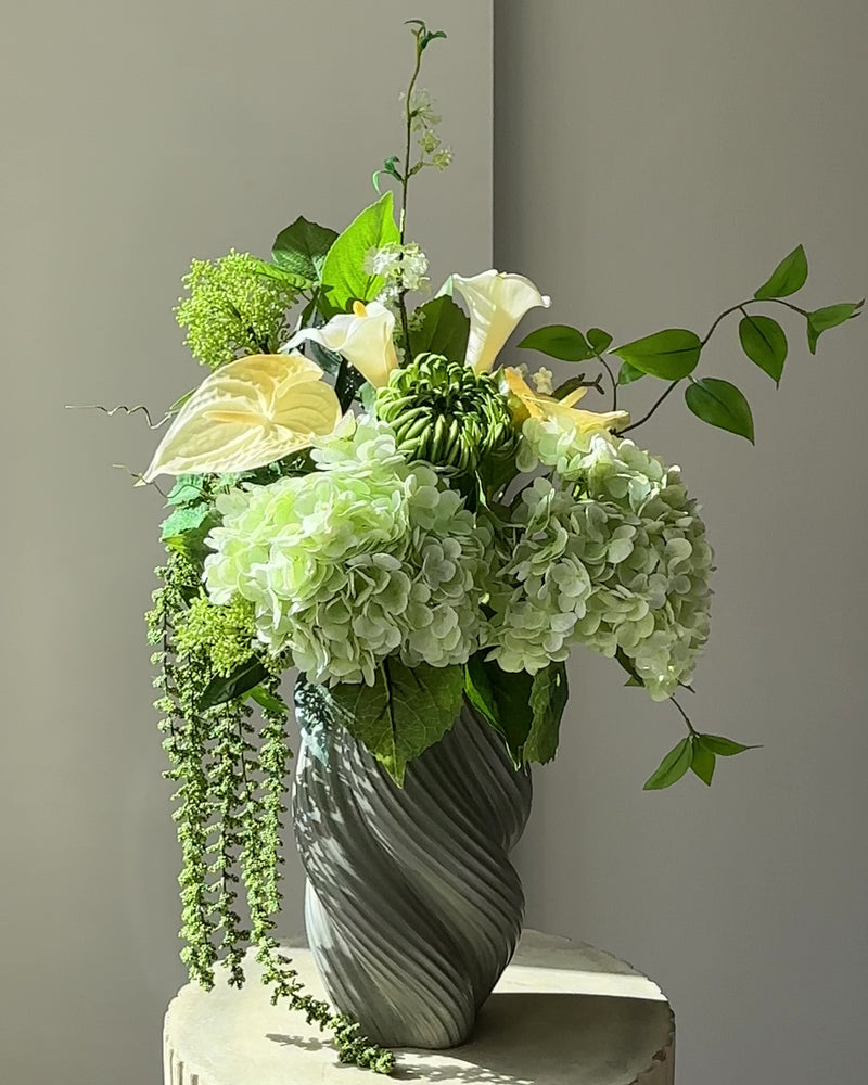 Green Atrium kunstig buket med grønne hortensiaer, hvide calla, grøn krysantemum og hængende amaranthus i en grågrøn snoet vase på en kannelleret stenpiedestal