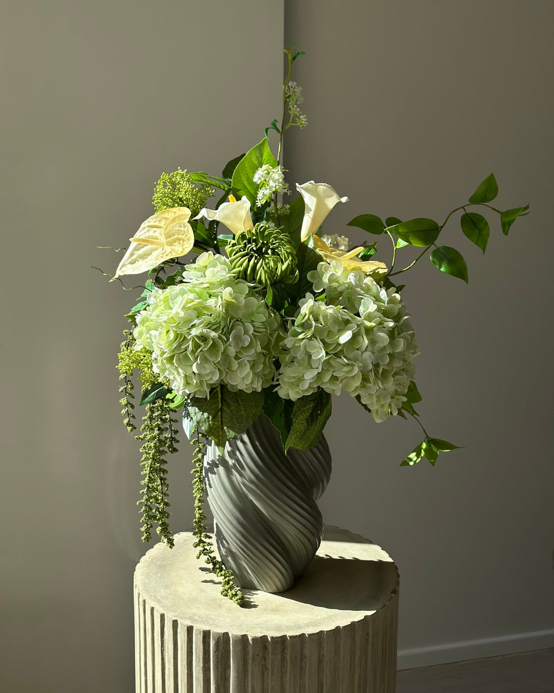 Green Atrium kunstig buket med grønne hortensiaer, hvide calla, grøn krysantemum og hængende amaranthus i en grågrøn snoet vase på en kannelleret stenpiedestal