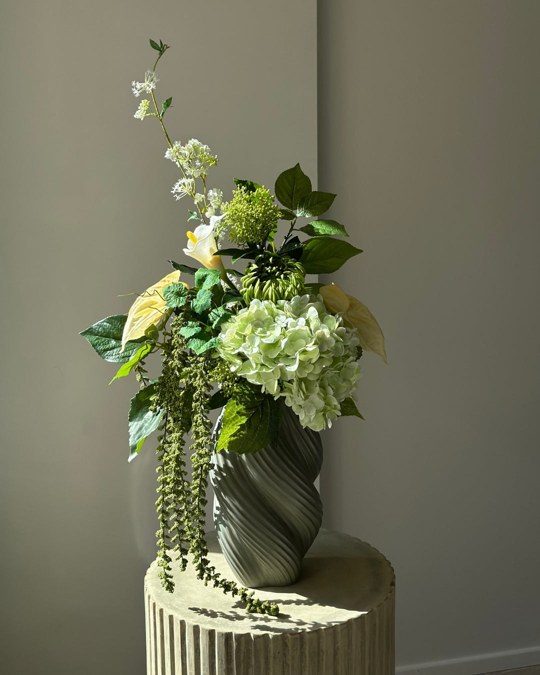 Green Loggia artificial bouquet with a light green hydrangea, a green cactus dahlia, a white calla lily and butter yellow anthurium in a grey-green twisted vase, with long green amaranthus trailing down onto a fluted stone pedestal